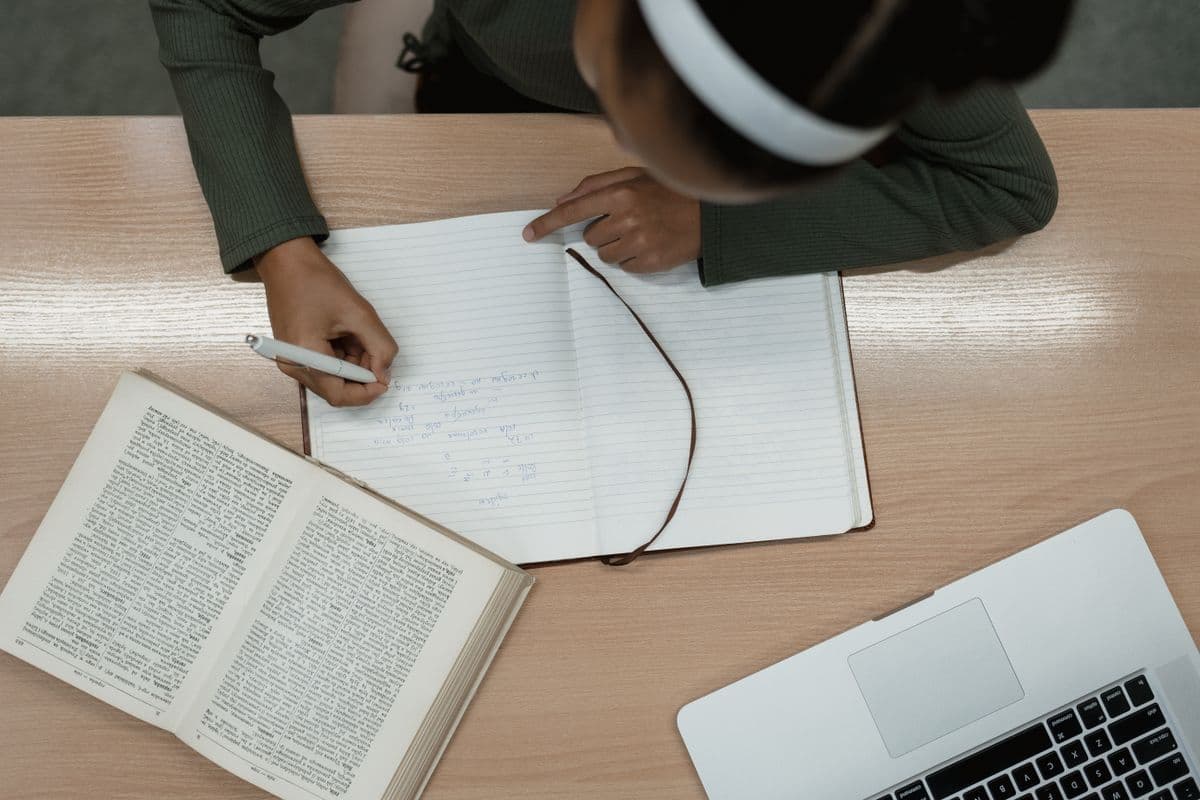 شخص يكتب ملاحظات في دفتر بجانب كتاب مفتوح وحاسوب محمول