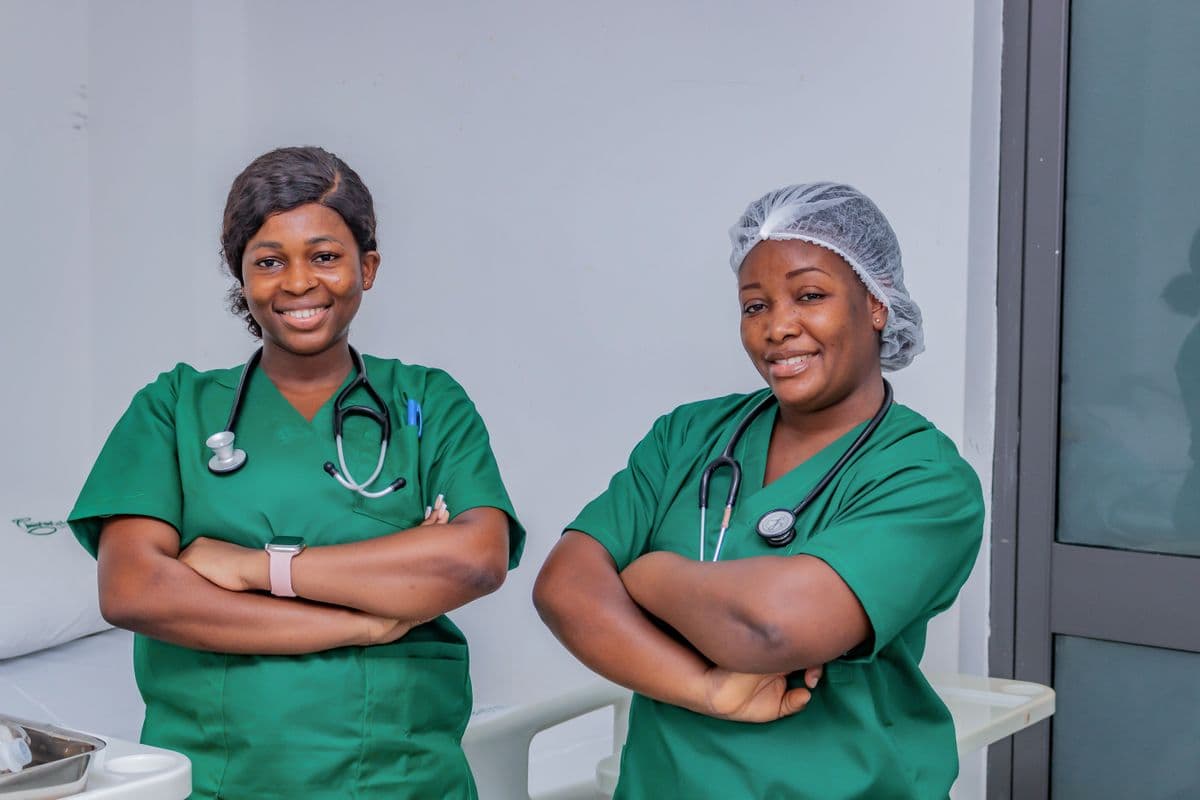 Two smiling nurses in green scrubs with stethoscopes and crossed arms in a hospital room