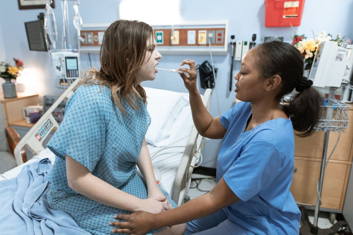 Nurse in blue scrubs taking a mouth swab from a patient sitting on a hospital bed