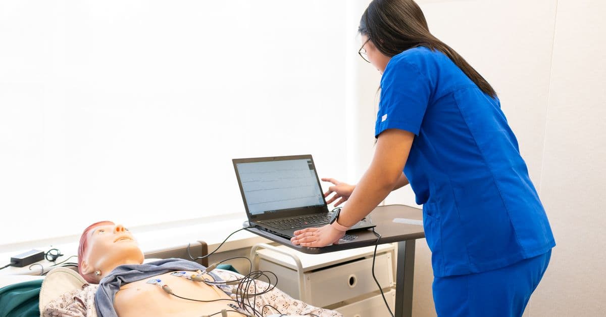 Nursing trainee in blue scrubs checks a laptop beside a patient manikin in a simulation lab
