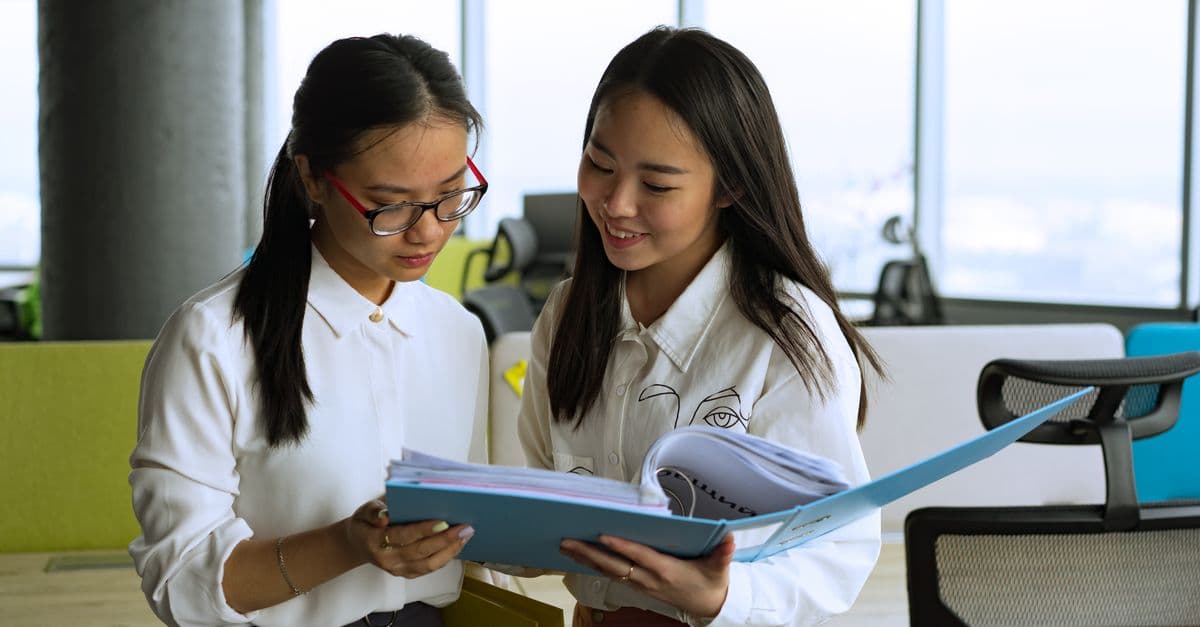 Two women standing in an office reading through papers in a blue folder together