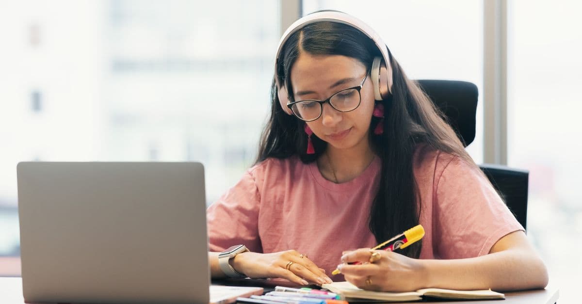 Young woman with headphones and glasses takes notes next to a laptop at a desk